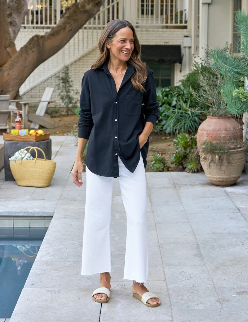 A woman with straight brown hair smiles on a patio, wearing the FRANK &amp; EILEEN Waverly NYC Button Up Shirt in Vintage Jersey British Royal Navy, paired with white pants. Plants and large pots decorate the background.
