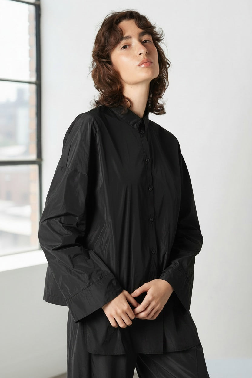 A person with wavy brown hair wears the KATHARINA HOVMAN - KIMONO BLOUSE by KATHARINA HOVMAN in dark gray, standing against a plain light background and looking slightly upward with a neutral expression.