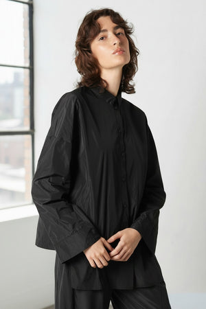 A person with wavy brown hair wears the KATHARINA HOVMAN - KIMONO BLOUSE by KATHARINA HOVMAN in dark gray, standing against a plain light background and looking slightly upward with a neutral expression.