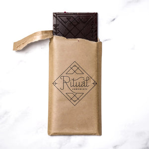 A rectangular chocolate bar with a pinkish wrapper adorned with minimalist illustrations of raspberries and abstract shapes. The packaging reads "RITUAL CHOCOLATE - THE APRES CHOCOLATE BAR 70%, Sparkling Wine Infused Chocolate with Dried Raspberries," 2.12 oz (60g).