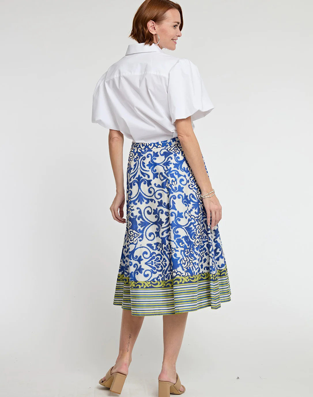 A person is modeling a white blouse with puffed sleeves and the HINSON WU - GLORIA SKIRT IN CERAMIC TILE PRINT, featuring a blue and white pattern with green and yellow striped accents at the hem. They are wearing beige open-toed, block-heeled sandals.