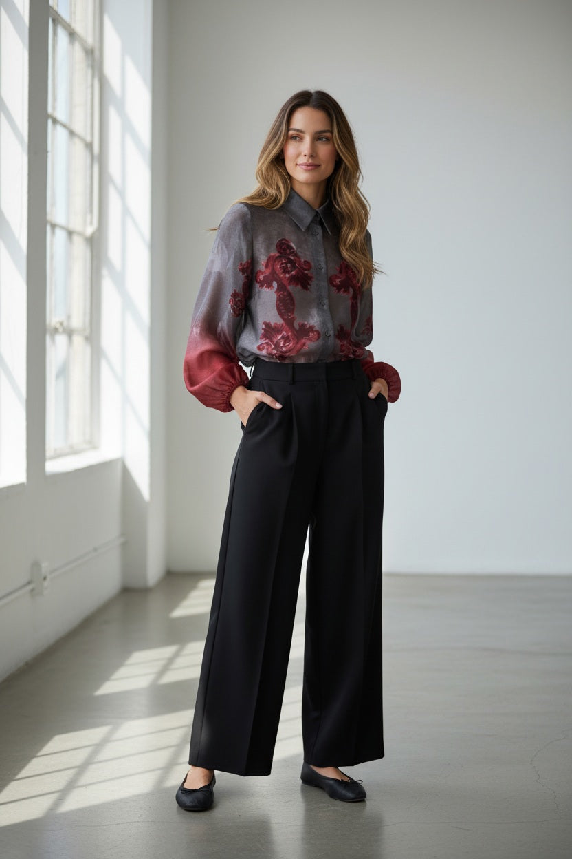 A person with short dark hair wears the MILVA MI - FLORAL RED BLOUSE by MILVA—featuring a gray and red floral pattern—and black high-waisted trousers, standing against a plain gray background.