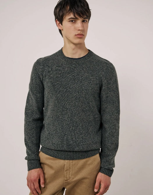 A young man with short dark hair wears the HARTFORD Seamless Wool Crew Pullover in Camo Green and tan pants, standing against a plain light-colored background, looking slightly to the side with a neutral expression.