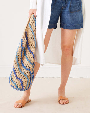 A person in white holds the MERSEA - CARLY CROCHET BOHO BAG by MERSEA, featuring large, colorful diagonal stripes of yellow, pink, red, green, and purple against a plain light background.