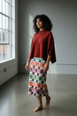 A woman stands indoors, wearing the BRITON COURT - SOPHIA OVERSIZED ONE SIZE SWEATER IN MULLBERRY by BRITON COURT, styled with a colorful plaid skirt. She smiles softly and touches her head with one hand, as natural light filters in from a window behind her.