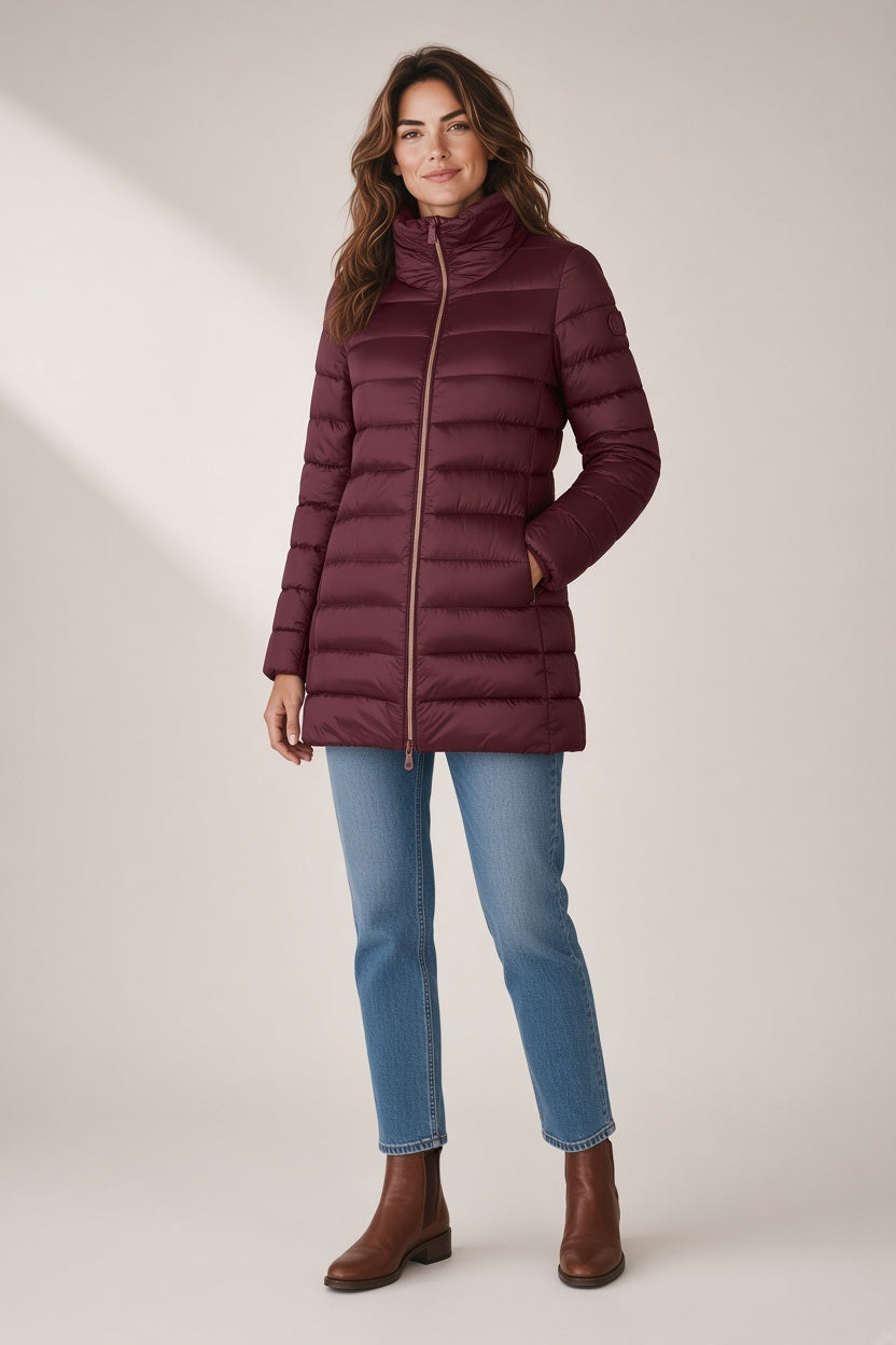 A woman wearing a knee-length, wine-colored SAVE THE DUCK - LYDIA PUFFER COAT IN WINE with animal-free Plumtech® padding, blue jeans, and brown leather ankle boots.