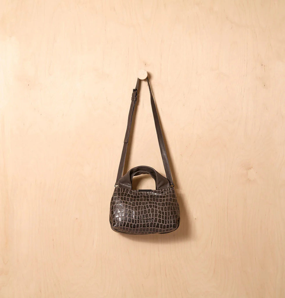 The DANIELLA LEHAVI Eveline Satchel in Grey Mosaic features a croco-inspired textured leather design with short handles and a long shoulder strap, epitomizing quiet luxury as it hangs gracefully against a wooden background.