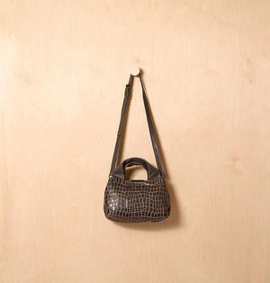 The DANIELLA LEHAVI Eveline Satchel in Grey Mosaic features a croco-inspired textured leather design with short handles and a long shoulder strap, epitomizing quiet luxury as it hangs gracefully against a wooden background.