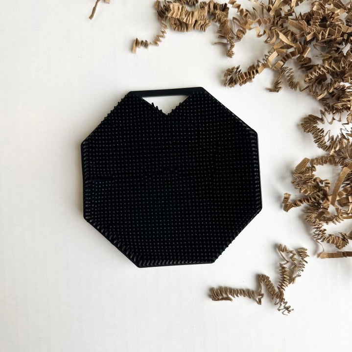 The SILICONE BODY AND EXFOLIATION BRUSH IN ONYX from SMILE BOUTIQUES features a black, octagonal silicone design with a textured, raised dot surface and a small triangular handle cutout at the top—perfect for use as a reusable shower brush or exfoliating body scrubber.