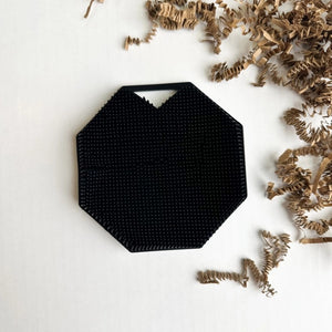 The SILICONE BODY AND EXFOLIATION BRUSH IN ONYX from SMILE BOUTIQUES features a black, octagonal silicone design with a textured, raised dot surface and a small triangular handle cutout at the top—perfect for use as a reusable shower brush or exfoliating body scrubber.