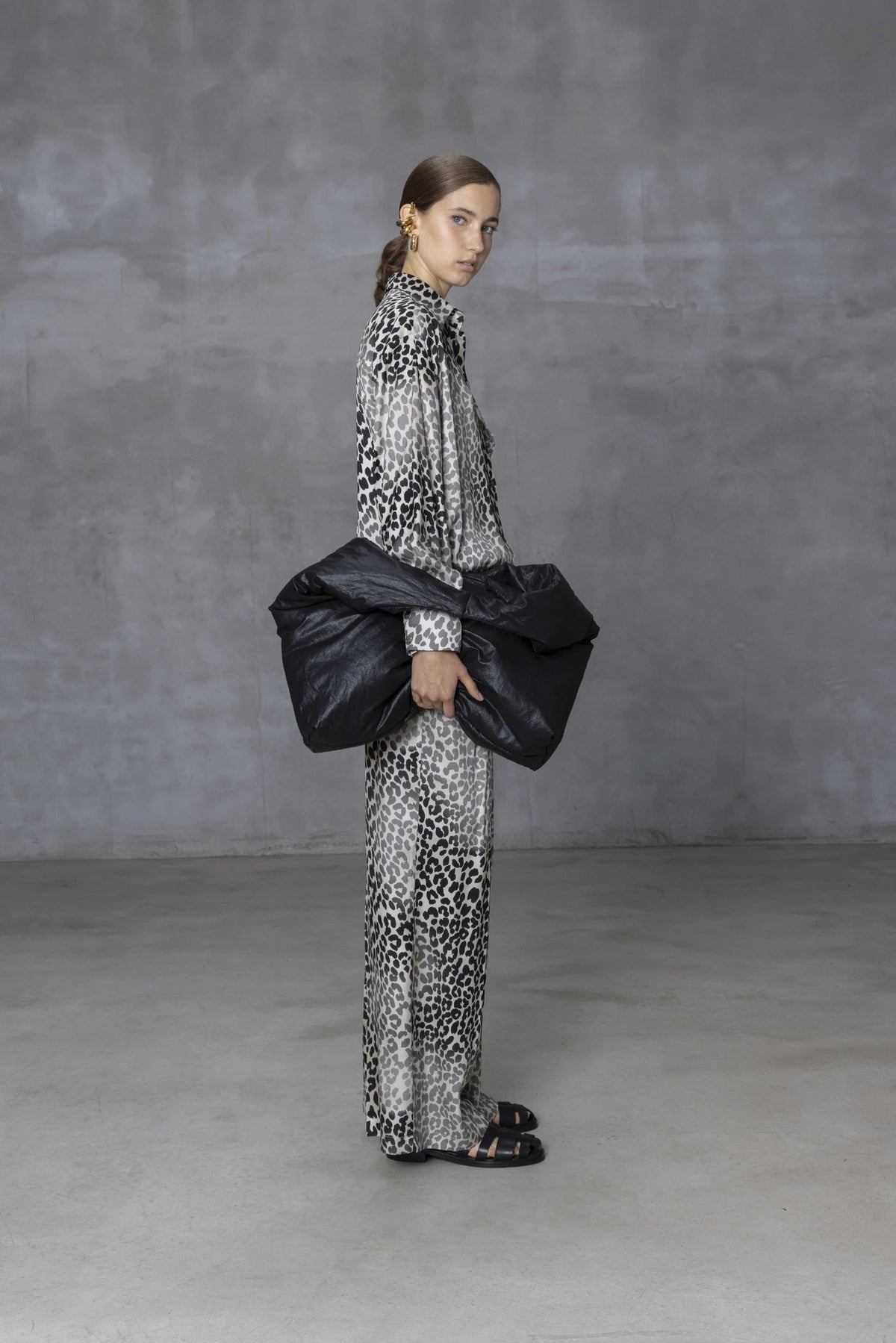A person in a long, leopard-print outfit stands against a gray wall, holding the NUDE - OVERSIZED HOBO BAG. Their hair is pulled back and they are wearing black sandals. The minimalist design of the concrete floor creates a neutral setting ideal for daily use.