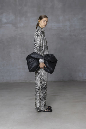 A person in a long, leopard-print outfit stands against a gray wall, holding the NUDE - OVERSIZED HOBO BAG. Their hair is pulled back and they are wearing black sandals. The minimalist design of the concrete floor creates a neutral setting ideal for daily use.