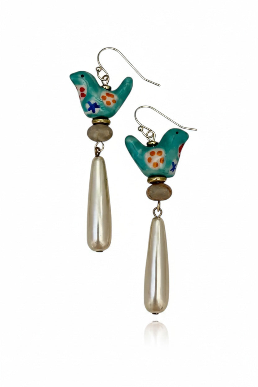 The SANDRINE FRANCE STUDIO - GEMMA EARRINGS feature turquoise ceramic bird charms with painted flowers, beige round beads, and long teardrop-shaped pearl dangles on gold hooks, shown against a white background.