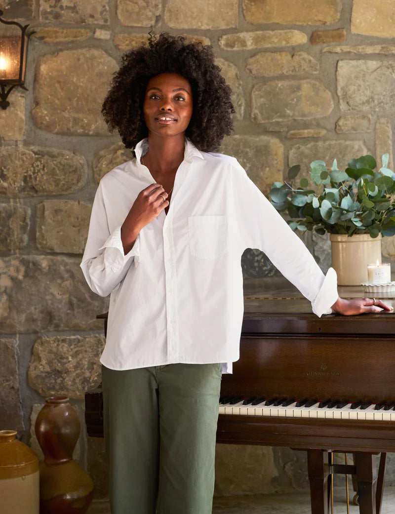 A person with curly hair, dressed in the FRANK & EILEEN OONA Continuous Sleeve Button Up Shirt in Superluxe Cotton White, and relaxed-fit green pants, stands facing away from the camera in front of a piano with their hands clasped behind their back. A stone wall and plants provide the backdrop.