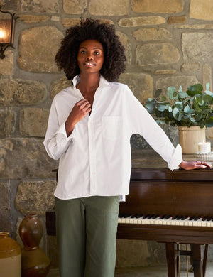 A person with curly hair, dressed in the FRANK & EILEEN OONA Continuous Sleeve Button Up Shirt in Superluxe Cotton White, and relaxed-fit green pants, stands facing away from the camera in front of a piano with their hands clasped behind their back. A stone wall and plants provide the backdrop.