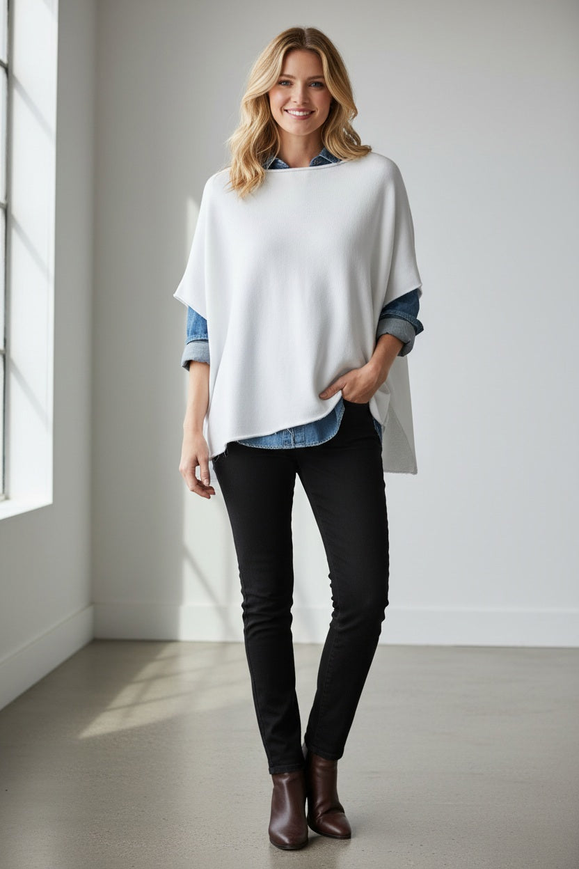 A woman with wavy brown hair stands near a window in a minimalist room, smiling softly with her hands by her sides. She is wearing the FRANK & EILEEN Olive Original Capelet in White—a loose, 100% cotton short-sleeve top—paired with blue jeans and black loafers.