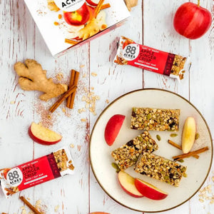 A package of 88 ACRES - APPLE GINGER CRISP SEED + OAT BAR rests on a light wooden surface. The primarily white and red wrapper emphasizes its nut-free, vegan, and gluten-free benefits. The bar is partially visible on the right side.