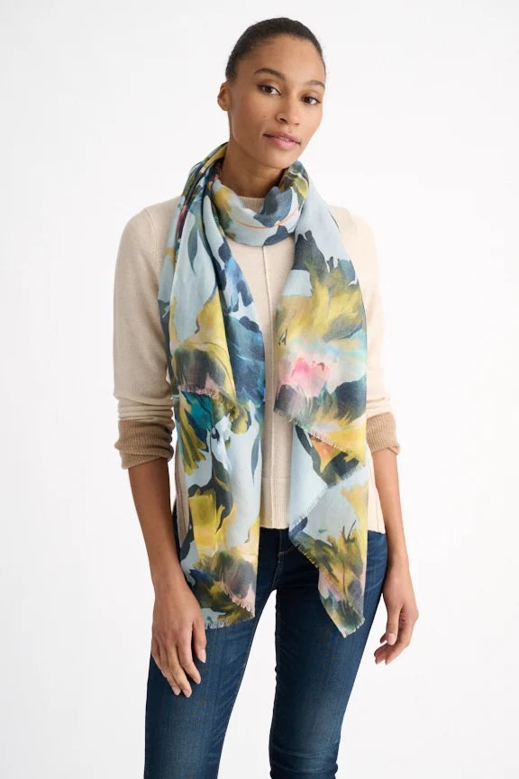 A woman wearing a beige sweater and jeans poses with the KINROSS CASHMERE Highland Blooms Scarf in Horizon—a long, lightweight floral print accessory from KINROSS—draped around her neck, standing against a plain white background.
