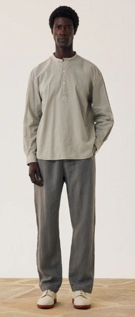 A man stands against a plain background wearing a light gray long-sleeve henley shirt, HARTFORD - LINEN JOGGY PANTS IN ARMY, and light-colored shoes, with his hands in his pockets.