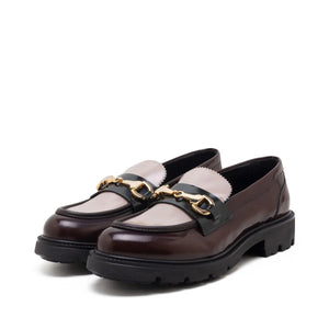 The MICHELE LOPRIORE - BRENDA TWO TONE LOAFER showcases a single dark brown leather design with a shiny finish. It features a black strap and gold-tone horsebit detailing across the top, complemented by a thick, chunky black sole for added durability. The shoe is set against a plain white background.