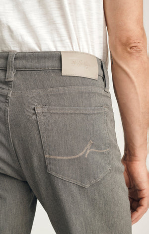 A man stands against a plain background wearing a dark brown sweater layered over a light collared shirt, paired with 34 HERITAGE - COURAGE STRAIGHT LEG PANTS IN STONE DIAGONAL by 34 HERITAGE, and brown shoes. One hand rests in his pocket and he maintains a neutral expression.