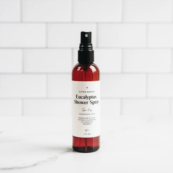A hand holds a brown spray bottle labeled “SPA DAY EUCALYPTUS SHOWER SPRAY” by GIFTEN MARKET—an uplifting eucalyptus aromatherapy mist—set against a black showerhead and white marble shower wall, ideal for transforming your shower into a spa-like escape at home.