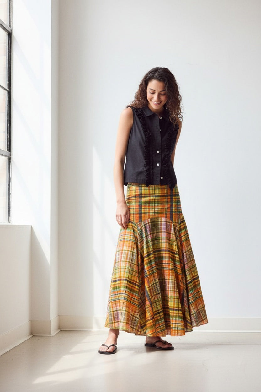 A woman stands indoors against a plain wall, wearing the HOPE FOR FLOWERS Circle Skirt in Spring Plaid, crafted from GOTS certified organic cotton lawn. She pairs it with a sleeveless black button-up top, black sandals, and has her hair tied back.