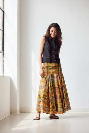 A woman stands indoors against a plain wall, wearing the HOPE FOR FLOWERS Circle Skirt in Spring Plaid, crafted from GOTS certified organic cotton lawn. She pairs it with a sleeveless black button-up top, black sandals, and has her hair tied back.