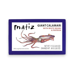 A bowl of rice salad with vegetables and wild-caught calamari sits beside a box labeled “Matiz España - Giant Calamari with White Beans and Paprika,” a small bowl of this gourmet seafood from Matiz España, a wooden fork and spoon, and a dish of green sauce infused with Spanish paprika on a white table.