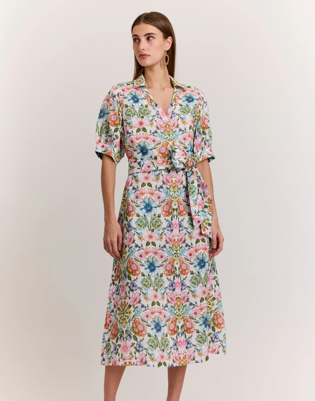 A woman wearing the HINSON WU Luca Elbow Sleeve Linen Blend Dress in Botanique, featuring a vibrant floral print, V-neck, elbow sleeves, and a tied waist belt, stands against a light background looking slightly to the side.