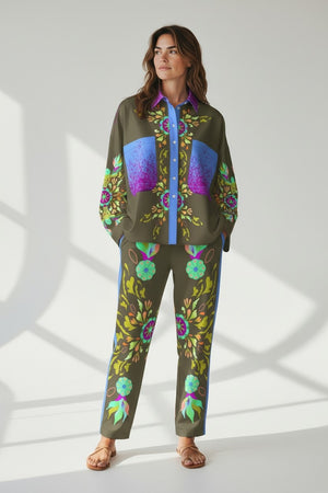 A person in FRANCO FERRARI SILK PRINT TROUSER—Italian-made, olive green with colorful florals and blue side stripes—matched with a coordinated top and tan sandals, stands against a bright background with soft shadows.