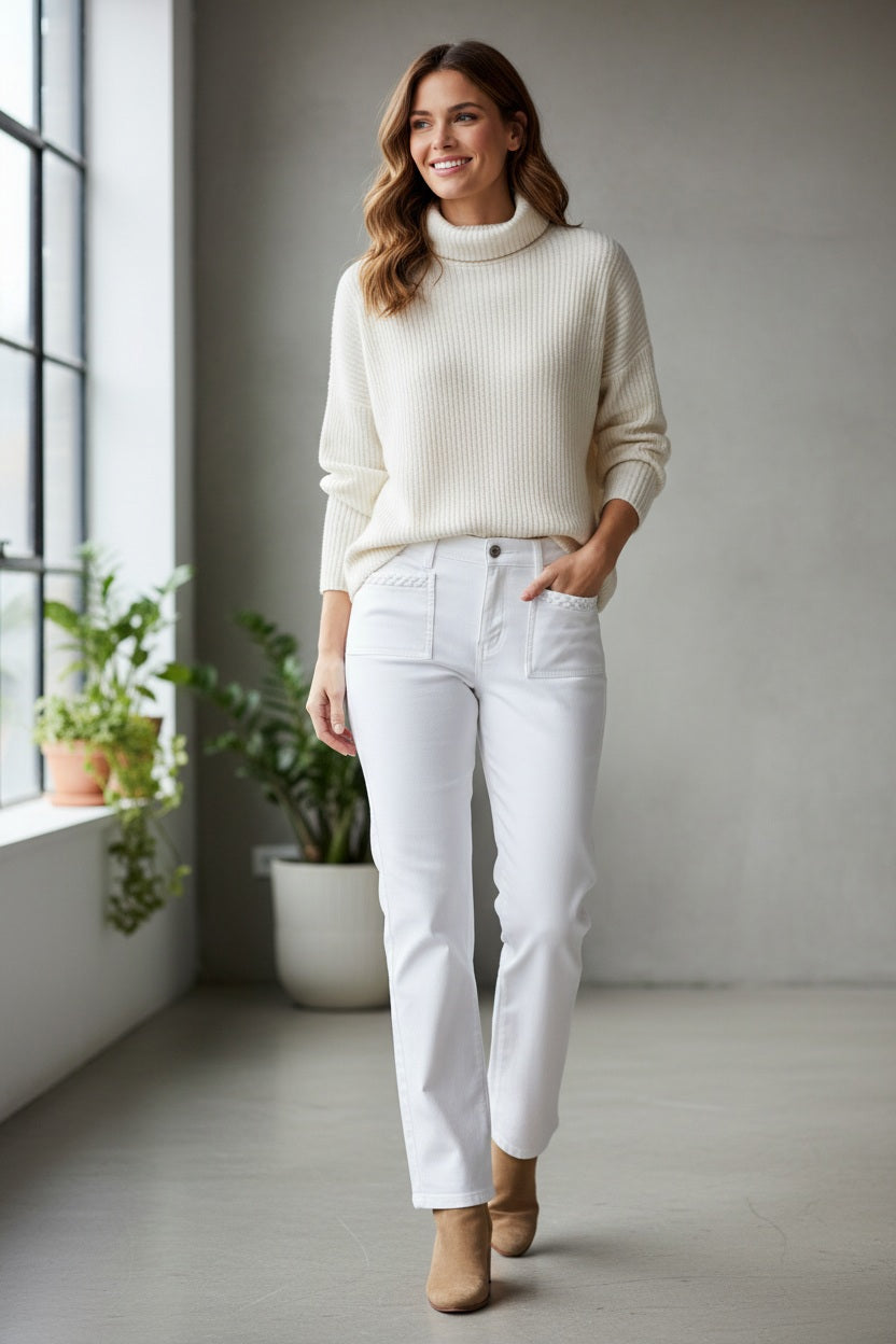 A woman stands indoors by a window, smiling as she wears the BRAX - MARY FIVE POCKET JEAN IN BOOTCUT WHITE from BRAX, styled with a cozy white sweater and tan ankle boots. Green potted plants are beside her and a neutral grey wall serves as the background.