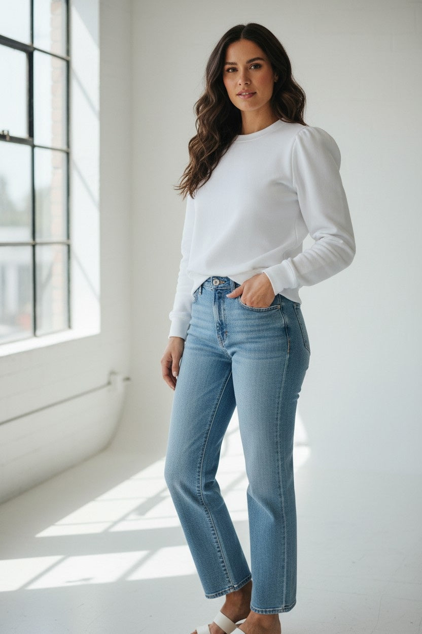 A woman stands in a bright room near large windows, wearing the KMJ - COOPER SWEATSHIRT IN WHITE and light blue jeans. She has long, wavy brown hair and poses with a relaxed expression.