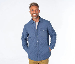 A man with a gray beard wears the CRIQUET - QUILTED SHACKET, a light blue piece with a pearl snap closure, along with a cream cap. He stands outdoors among pine trees and greenery in a forested area.