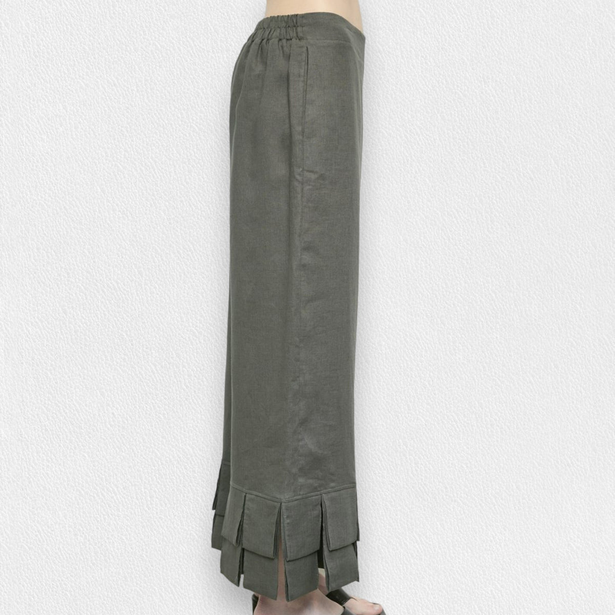A person is wearing MEIMEIJ&#39;s olive green linen pull-on pants with a pleated bottom design and ruffle cuffs. The pants reach down to their ankles, and the background is a textured white wall. Only the lower half of the body is visible.