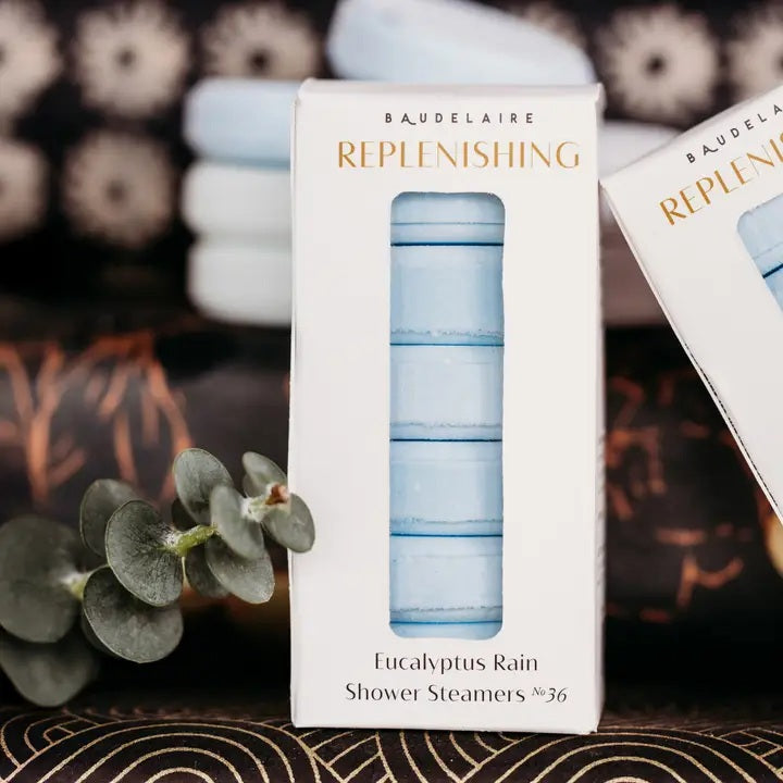 A white box labeled “BAUDELAIRE - REPLENISH EUCALYPTUS RAIN SHOWER STEAMER” by BAUDELAIRE, featuring a window that displays stacked round blue and white shower steamers inside, provides a refreshing aromatherapy experience with the invigorating scent of eucalyptus.