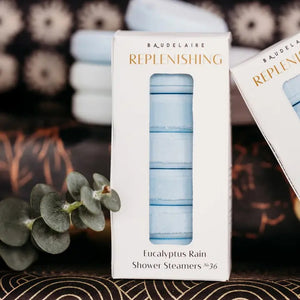 A white box labeled “BAUDELAIRE - REPLENISH EUCALYPTUS RAIN SHOWER STEAMER” by BAUDELAIRE, featuring a window that displays stacked round blue and white shower steamers inside, provides a refreshing aromatherapy experience with the invigorating scent of eucalyptus.