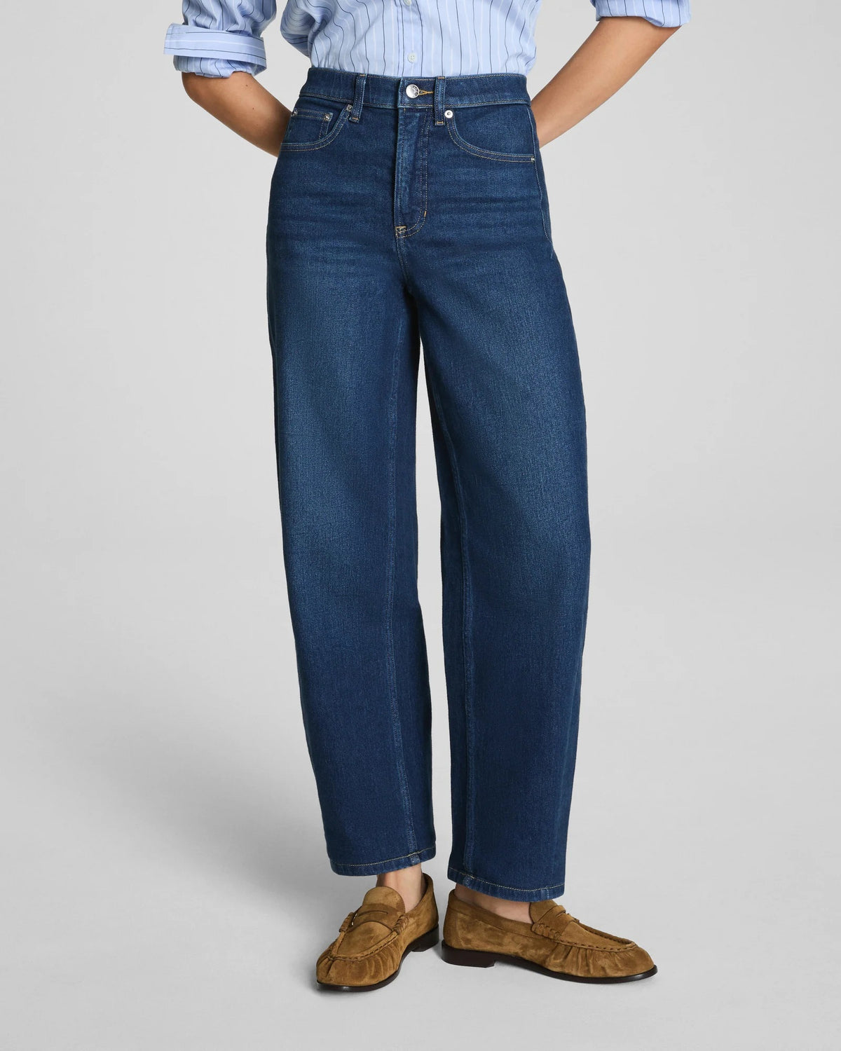 Here is the rewritten sentence using your product data: A woman with long dark hair and a relaxed expression stands against a plain background wearing the SPANX SPANXSHAPE Authentic 360 Barrel Leg Jean in Adriatic Blue, paired with a matching blue denim jacket, light blue shirt, and brown suede loafers.