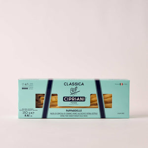 A rectangular box of Cipriani Organic Egg Pappardelle Pasta, produced by Atalanta Corporation and made in Italy, is displayed against a pale background. The light blue packaging with dark blue and white accents features branding details, cooking information, and a small Italian flag denoting its origin.