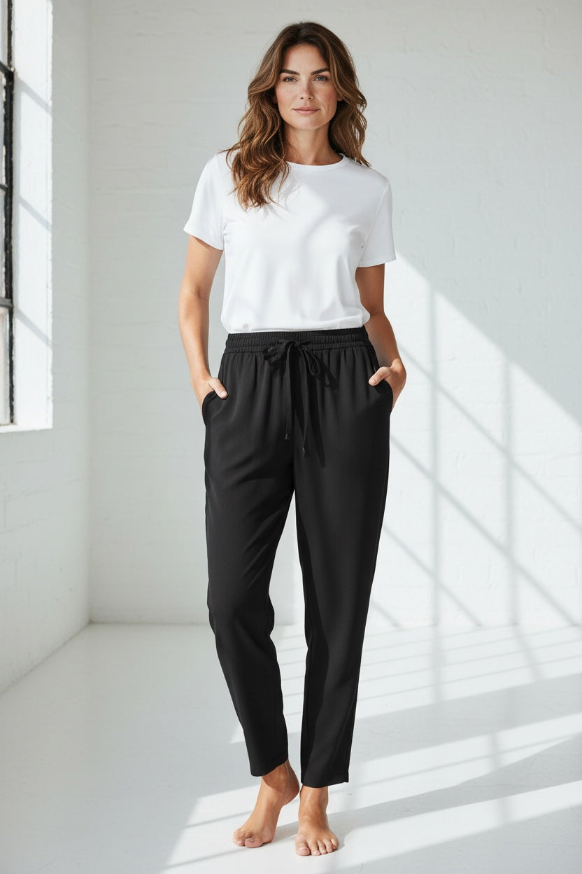 A person wears the MEIMEIJ DRAWSTRING TROUSER PANT—high-waisted, loose-fitting black pants in soft polyester with a tie belt—standing barefoot against a light textured background.