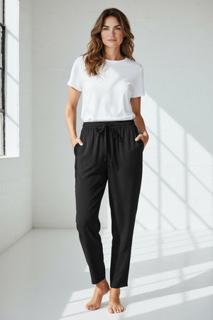 A person wears the MEIMEIJ DRAWSTRING TROUSER PANT—high-waisted, loose-fitting black pants in soft polyester with a tie belt—standing barefoot against a light textured background.