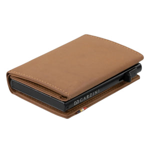 The GARZINI - SLIM TRIFOLD RFID-blocking leather wallet in camel brown is shown open, displaying a blue card, a white card, and a large “NO CASH VALUE” play bill. “GARZINI” is embossed on the left side. Includes metal card holder and coin pocket.