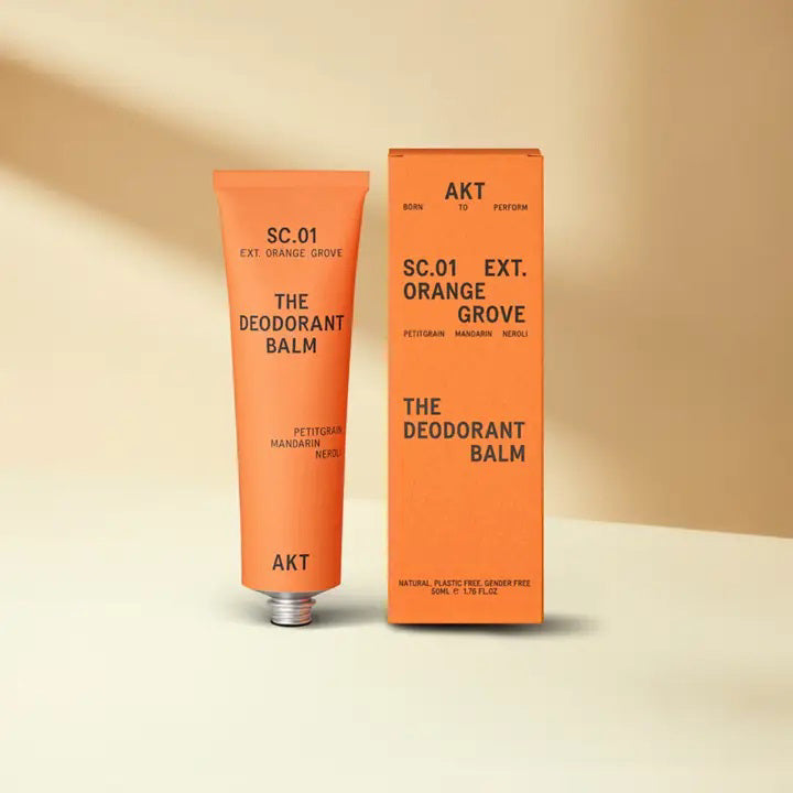 An orange tube and box of AKT LONDON’s DEODORANT BALM ORANGE GROVE—an eco-friendly, aluminum-free deodorant with petitgrain, mandarin, and neroli—are shown on a beige surface against a soft cream background.