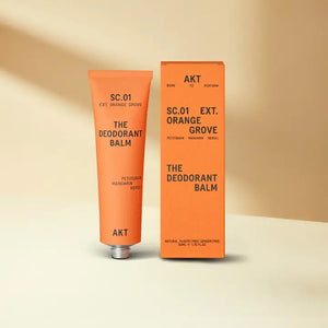 An orange tube and box of AKT LONDON’s DEODORANT BALM ORANGE GROVE—an eco-friendly, aluminum-free deodorant with petitgrain, mandarin, and neroli—are shown on a beige surface against a soft cream background.