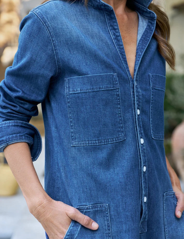 A woman smiles while standing outdoors, wearing the FRANK & EILEEN Southern Ireland Utility Dress in Italian Dream Light Denim 1984 wash, featuring rolled-up sleeves and six pockets where she rests her hands. Behind her, a basket and fruit are arranged on a table near greenery.