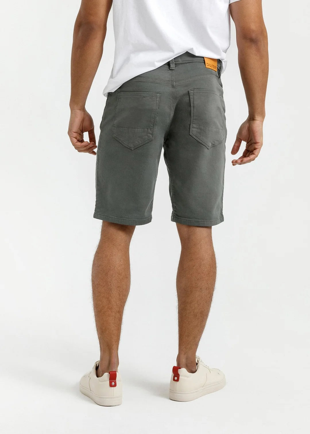 A person stands against a plain light background, wearing a white T-shirt, DUER No Sweat Relaxed Shorts in Gull made from stretch fabric, and white sneakers, with one hand in their pocket.