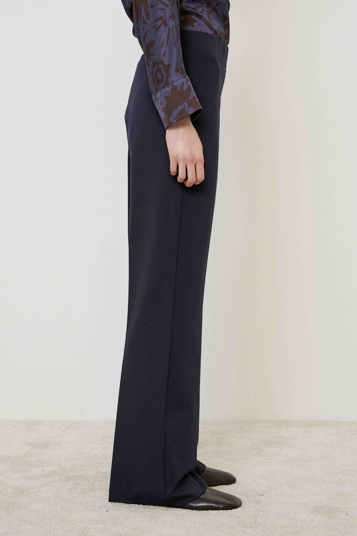 A person in MEIMEIJ PULL ON PONTE TROUSER PANT, a patterned long-sleeve blouse, and black shoes stands on a light carpet against a plain wall, highlighting this versatile MEIMEIJ wardrobe essential with a tailored look.