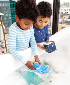 The MUSEE - YOU’RE OUT OF THIS WORLD BATH BALM from MUSEE rests next to its vibrant space-themed box, which features a smiling cartoon astronaut and a rocket on top, highlighting the playful appeal of natural bath products.