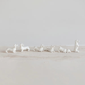 Six small white STONEWARE DOGS by CREATIVE COOP, each in a unique pose, are displayed in front of a six-compartment box, with each compartment holding a decorative dog figurine.