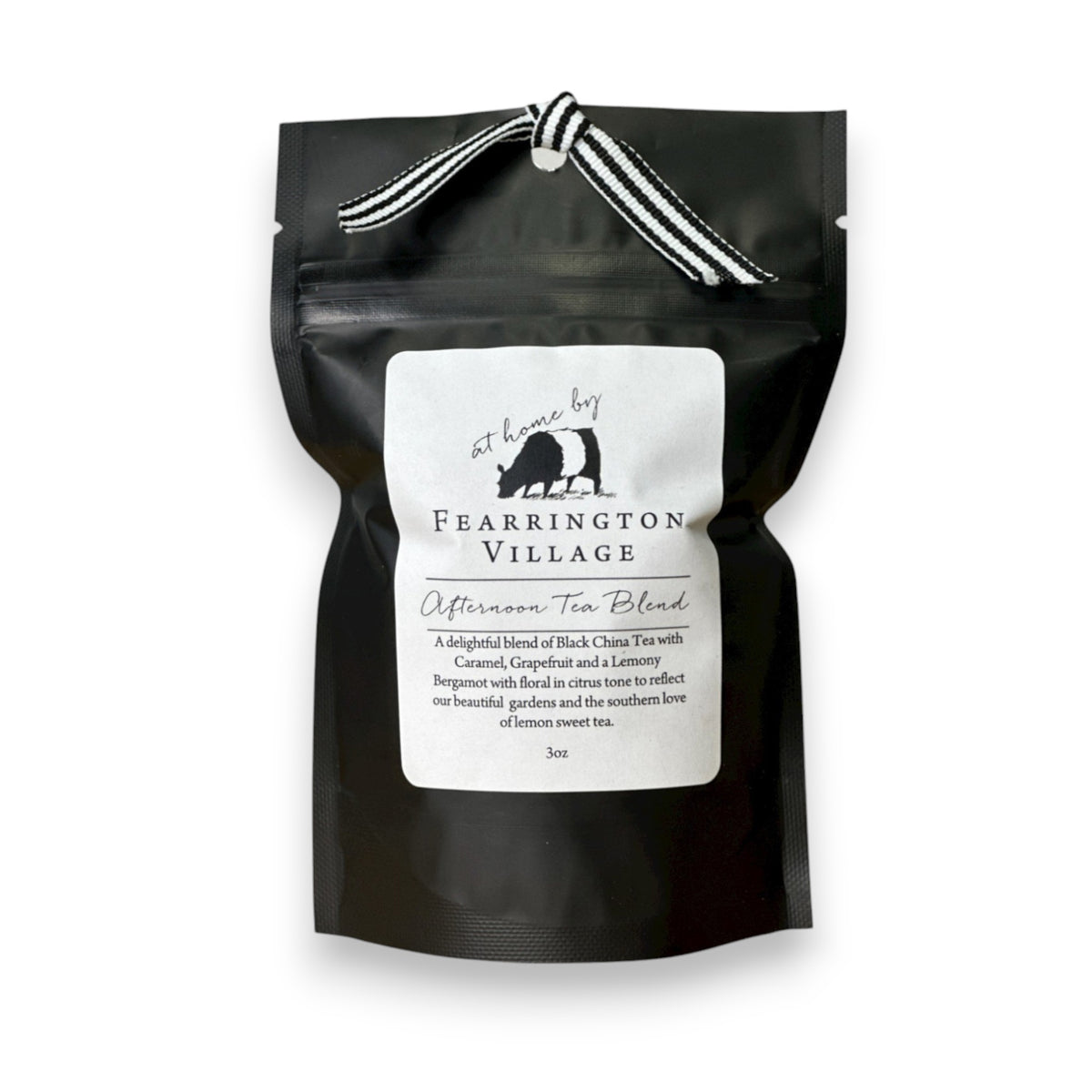 A black foil tea bag labeled &quot;FEARRINGTON SIGNATURE COLLECTION - FEARRINGTON SIGNATURE AFTERNOON TEA BLEND&quot; from Fearrington House, featuring a small white label with an image of a bear holding a bird. The bag contains loose leaf tea and is tied at the top with a black and white striped ribbon. It rests on a light gray surface.
